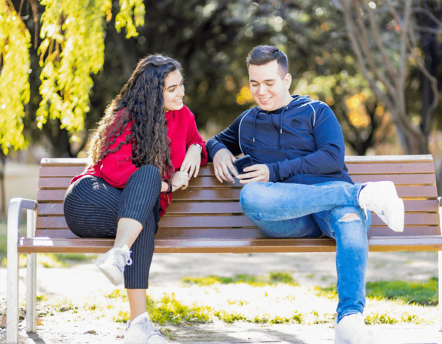 Two young adults sit on a bench in a park, chatting and looking at a phone.