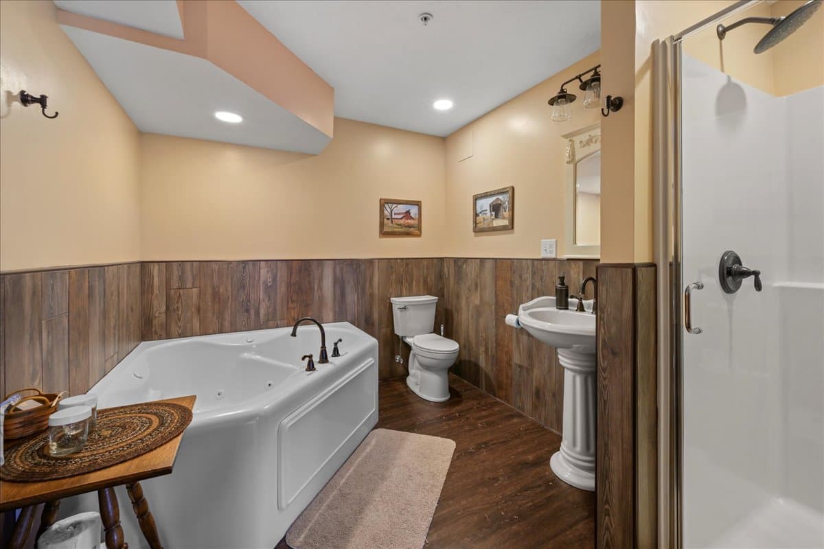 A cozy bathroom featuring a large jacuzzi tub, toilet, pedestal sink, and a glass shower, with warm-toned walls and wood accents.