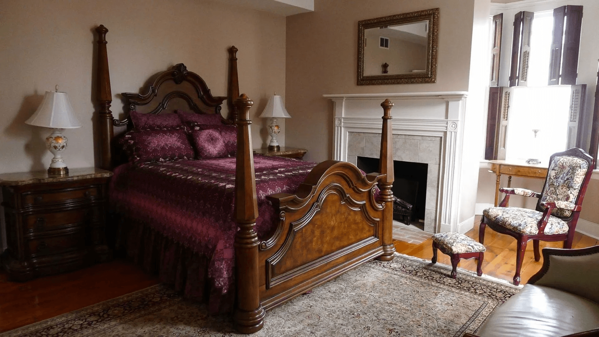 A warmly decorated bedroom featuring a four-poster bed, side table with lamp, and floral accent chair near a fireplace.